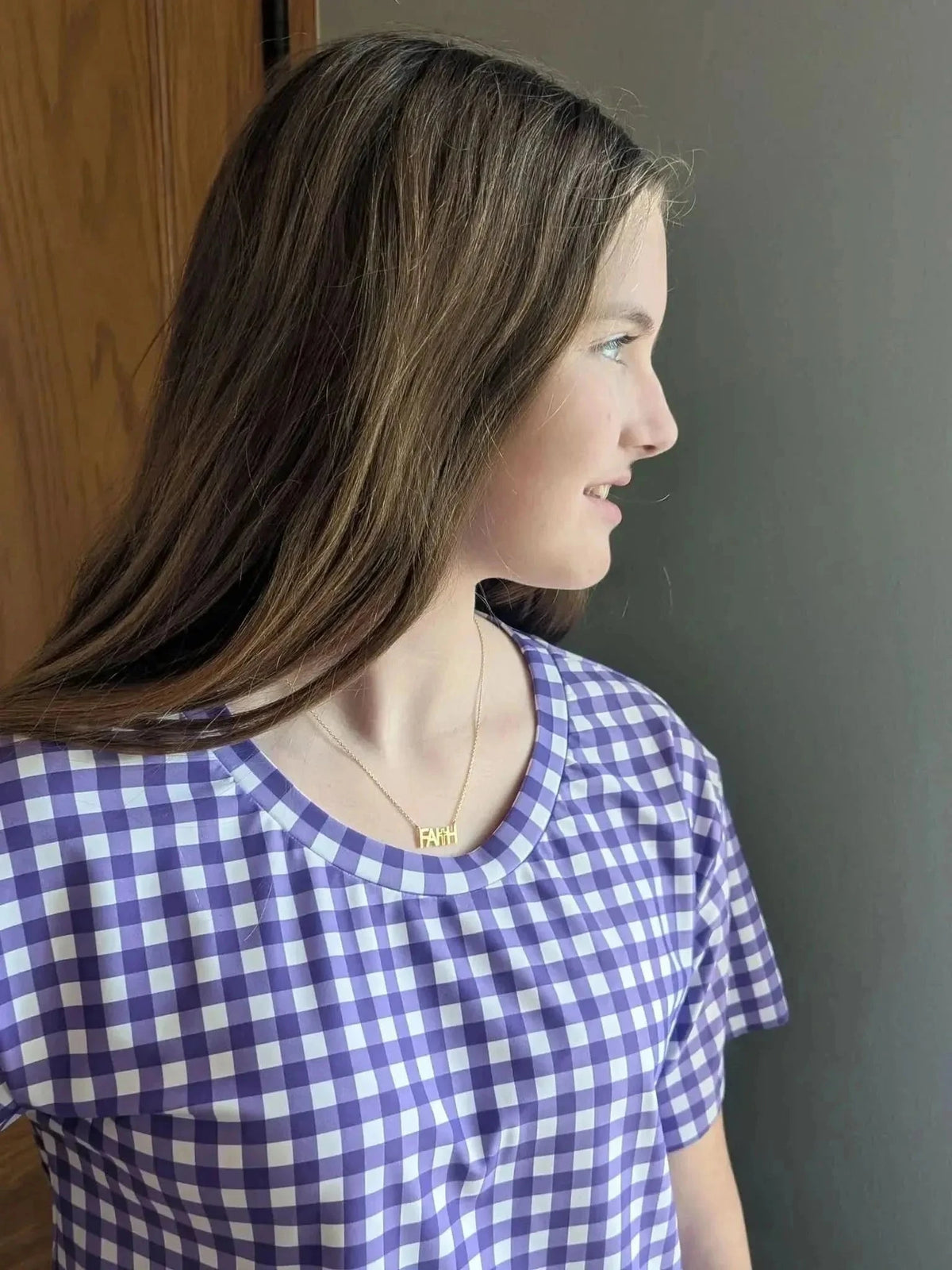 A young woman wears a purple and white gingham shirt with a "FAITH" necklace.