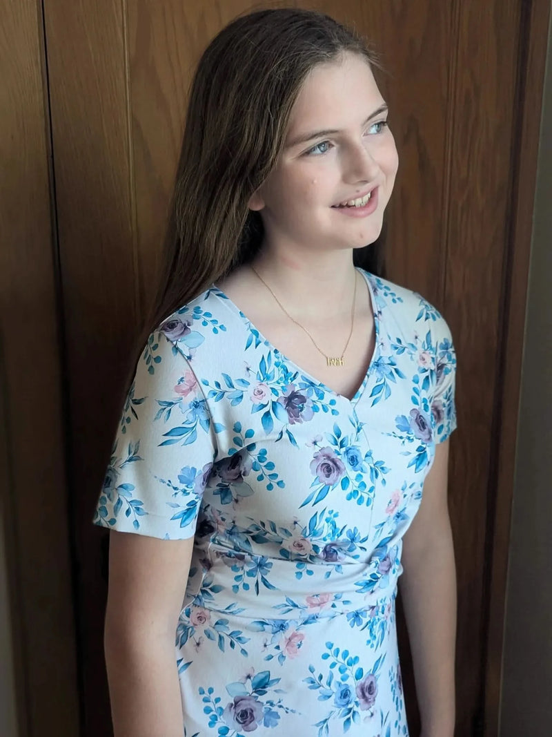 A young girl wears a blue watercolor floral dress with a gold necklace.