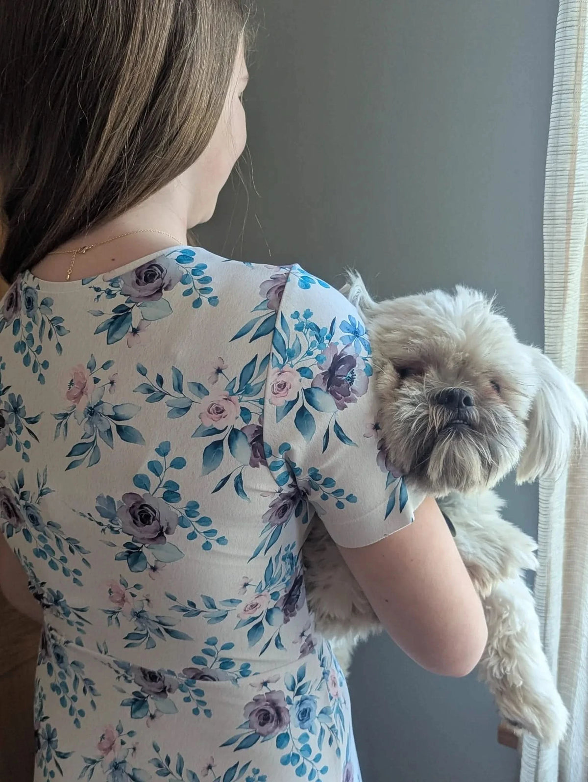 A woman in a blue watercolor floral dress holds a small white dog.