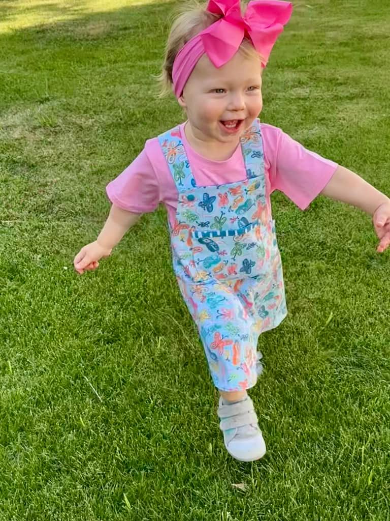 A child in a butterfly-patterned overall dress and pink shirt runs on grass.