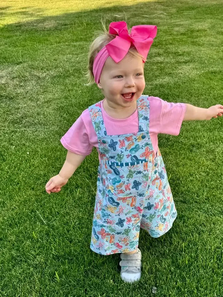 A smiling toddler wears a pink shirt and a butterfly-patterned overall dress.