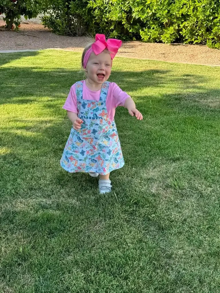 A toddler in a butterfly-patterned dress and pink shirt smiles on a grassy lawn.