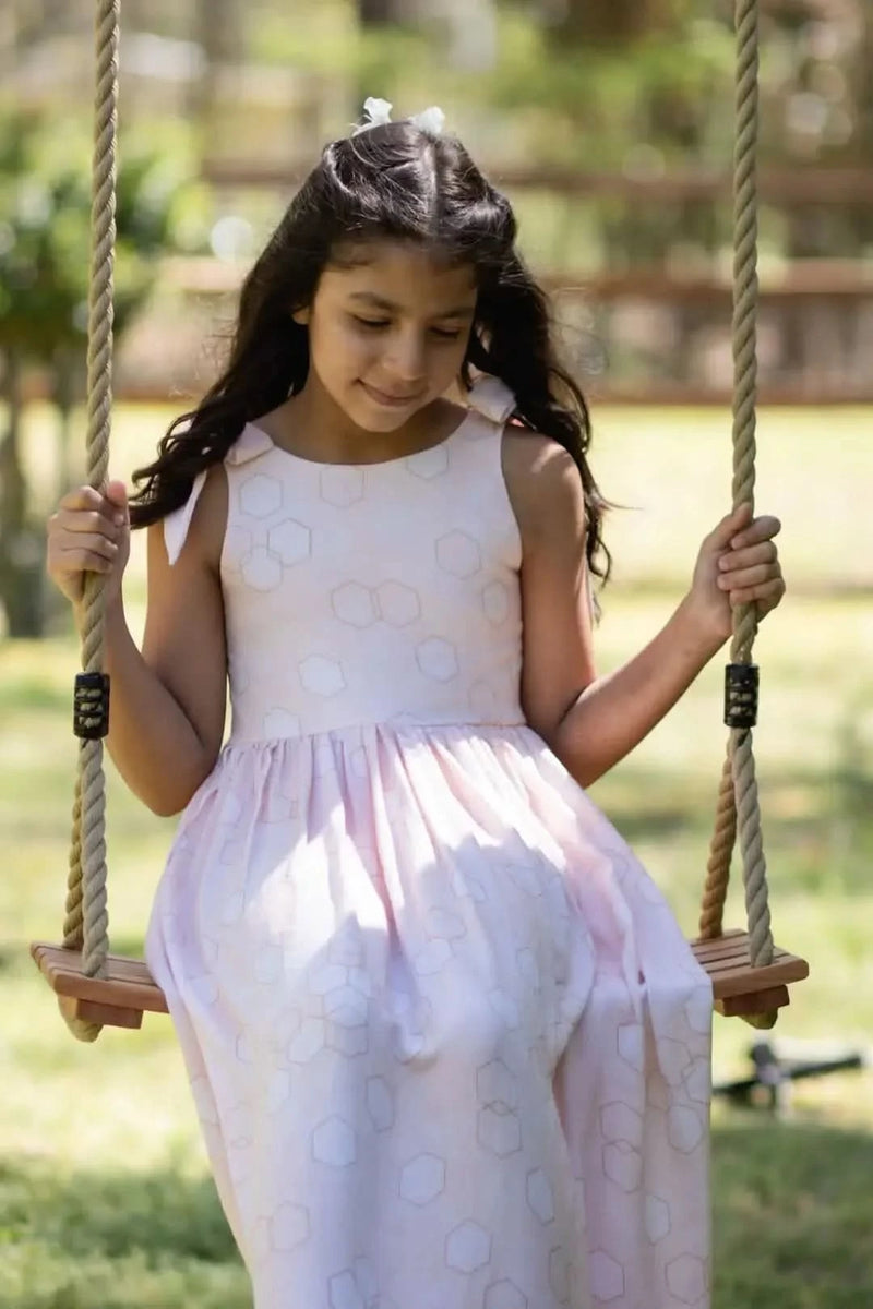 A young girl in a light pink dress with a subtle geometric pattern sits on a swing.
