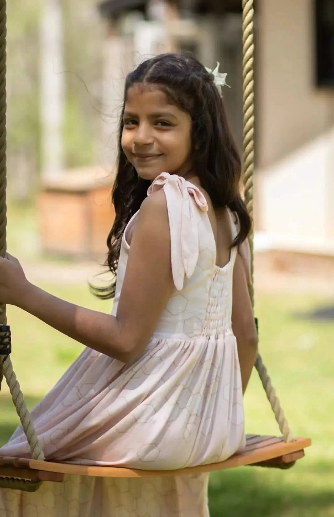 A young girl in a peachy geometric print dress sits on a wooden swing.
