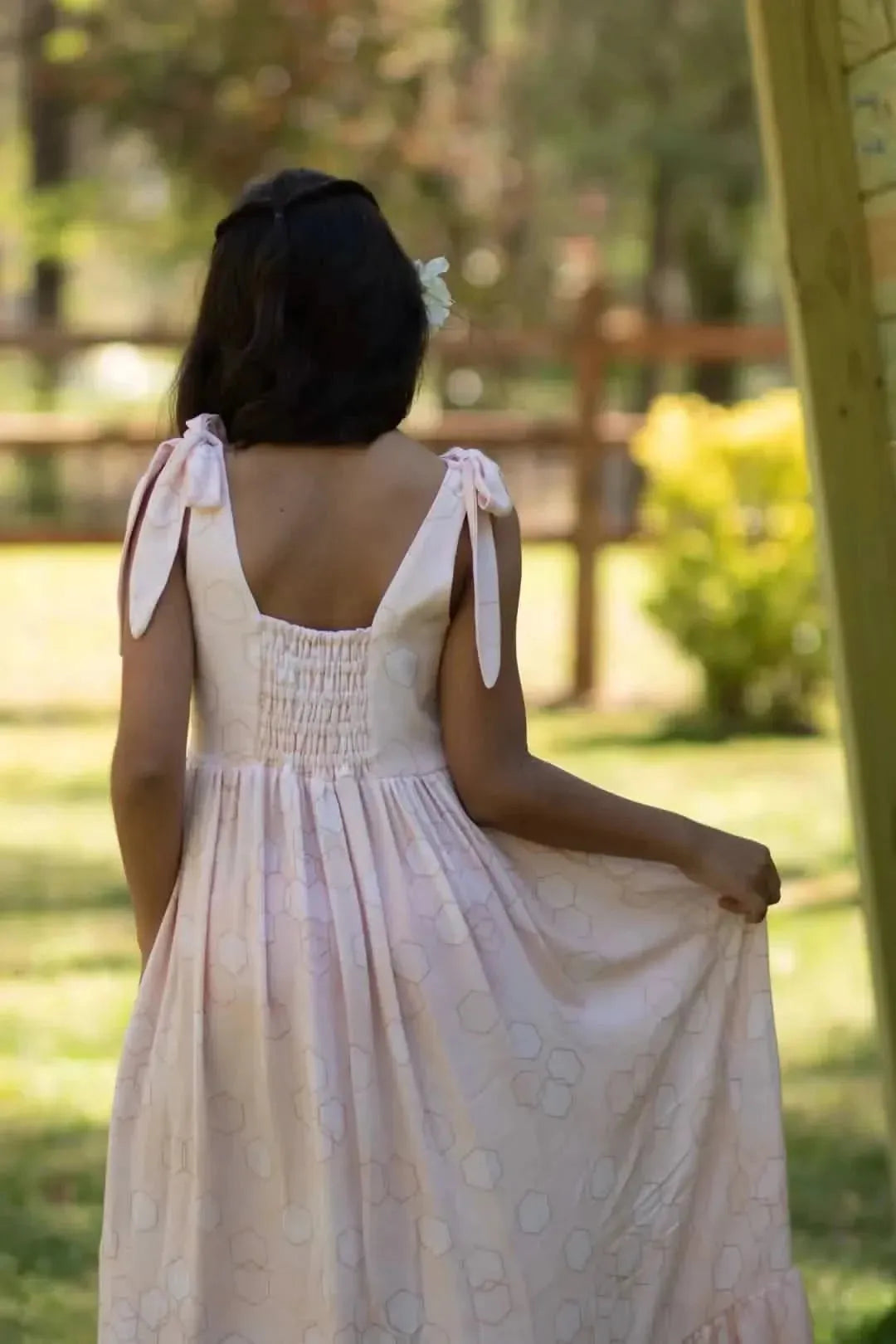 Back view of a woman in a pink hexagon-print dress with shoulder ties in a garden.