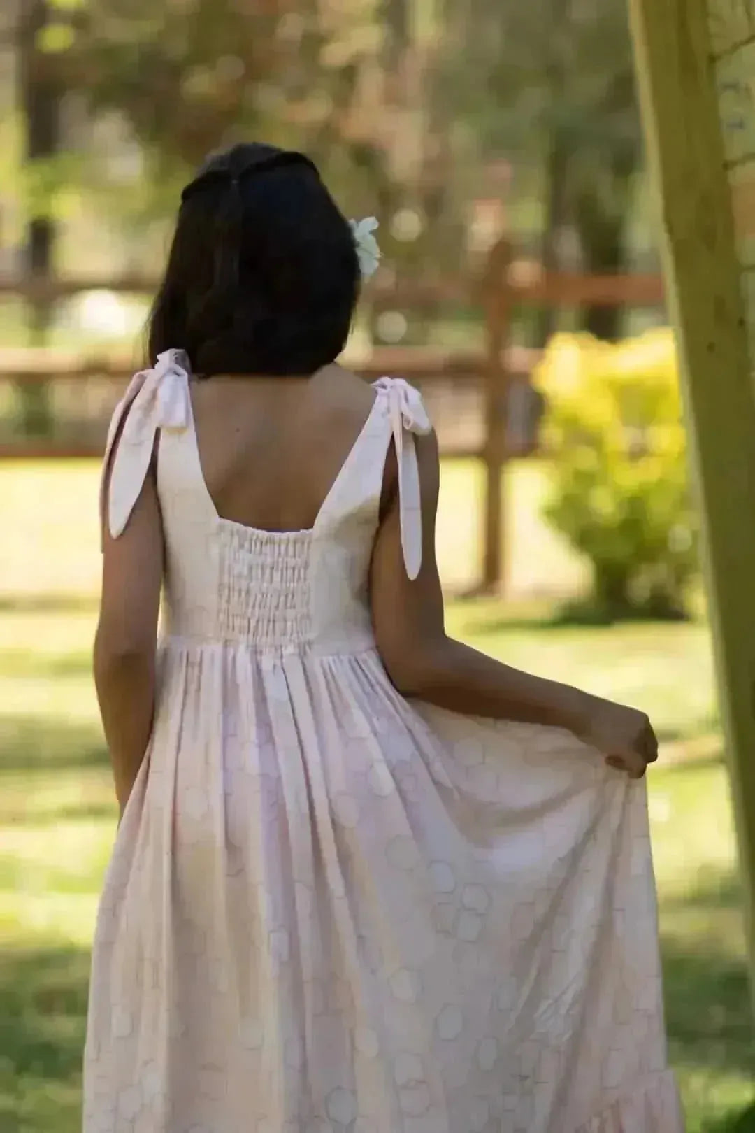 Back view of a woman in a pink hexagon-print dress with shoulder ties in a garden.