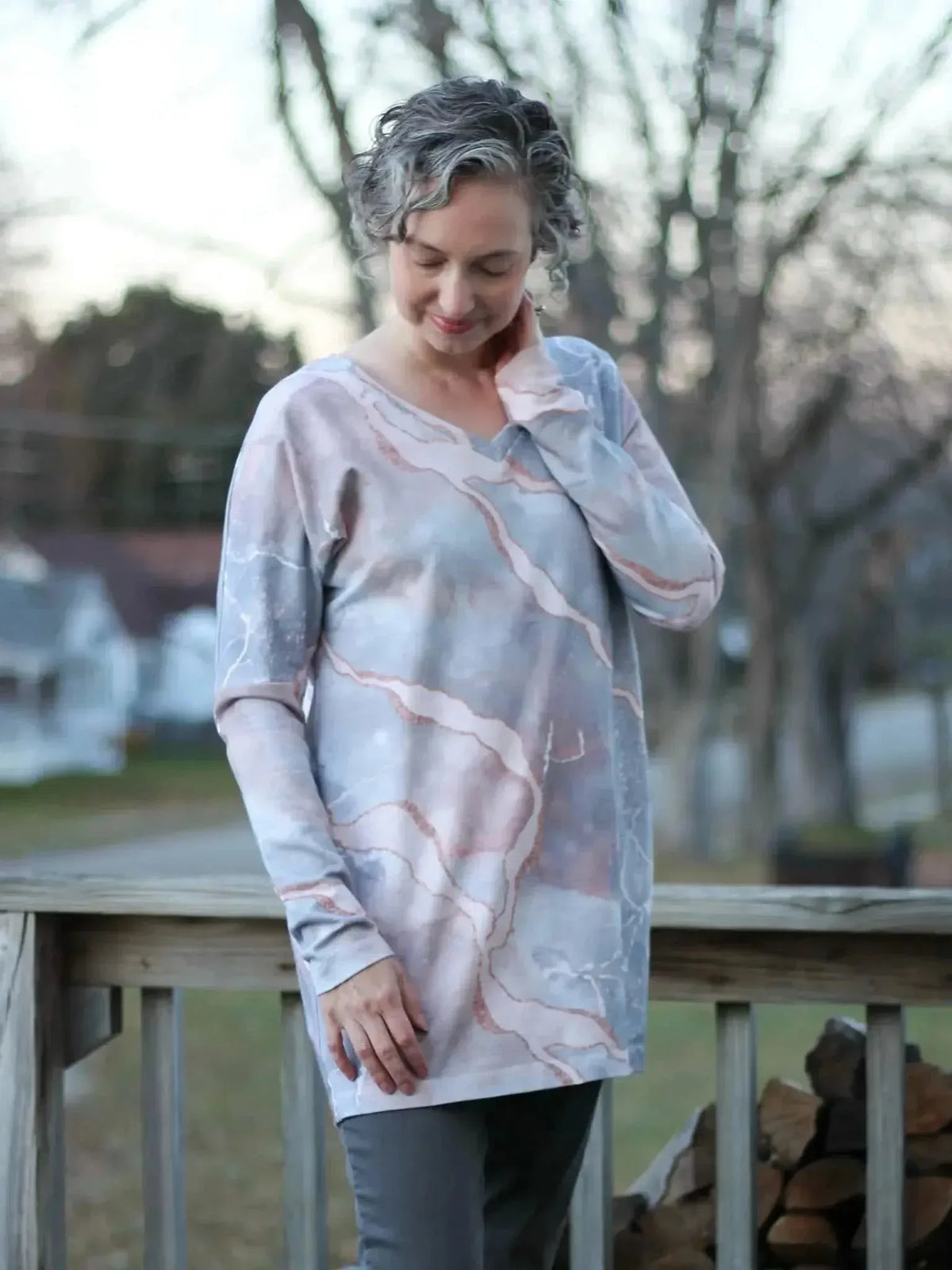 Woman modeling a long-sleeved, V-neck top in pink marble fabric on a porch.