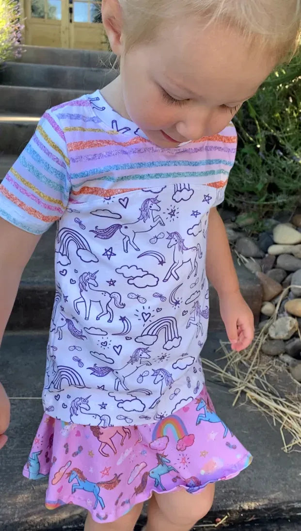 A child wears a white shirt with purple unicorn outlines and a pink unicorn skirt.