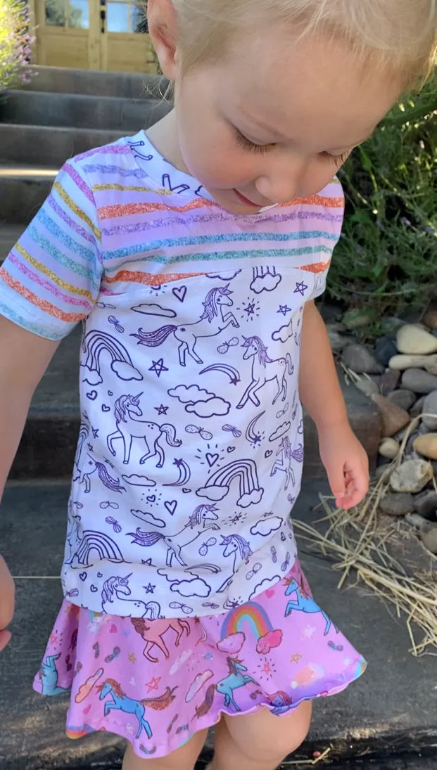 A child wears a white shirt with purple outline unicorns and a pink unicorn skirt.