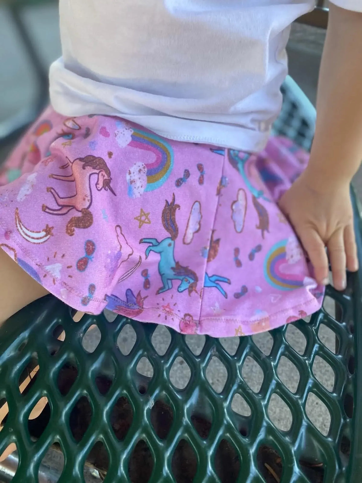 A child wears a pink skirt with unicorns, rainbows, and sprinkles, sitting on a green metal bench.