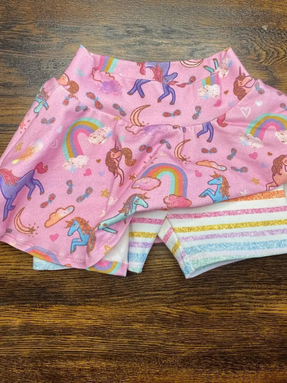 A pink unicorn sparkle skirt with rainbow striped shorts is displayed on a wooden surface.