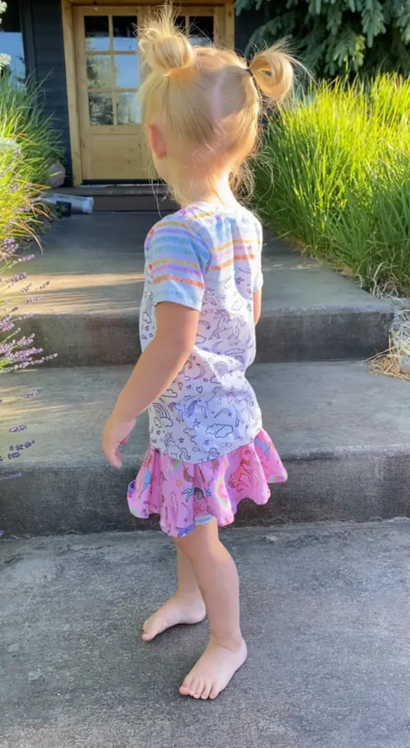 A young girl from behind wears a pink unicorn skirt and a unicorn and rainbow shirt.