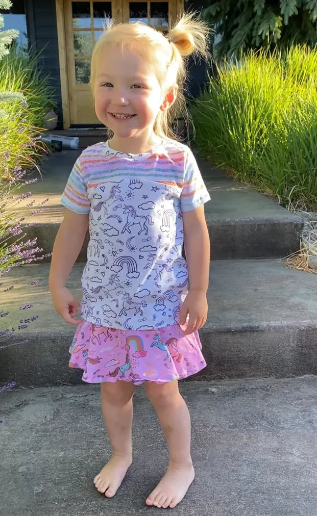 A young child smiles, wearing a unicorn-patterned shirt and a pink unicorn skirt.