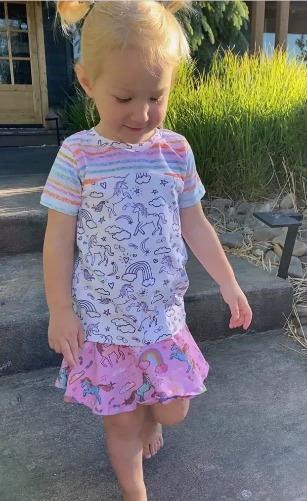 A young child wears a pink unicorn sparkle skirt and a unicorn-patterned shirt.