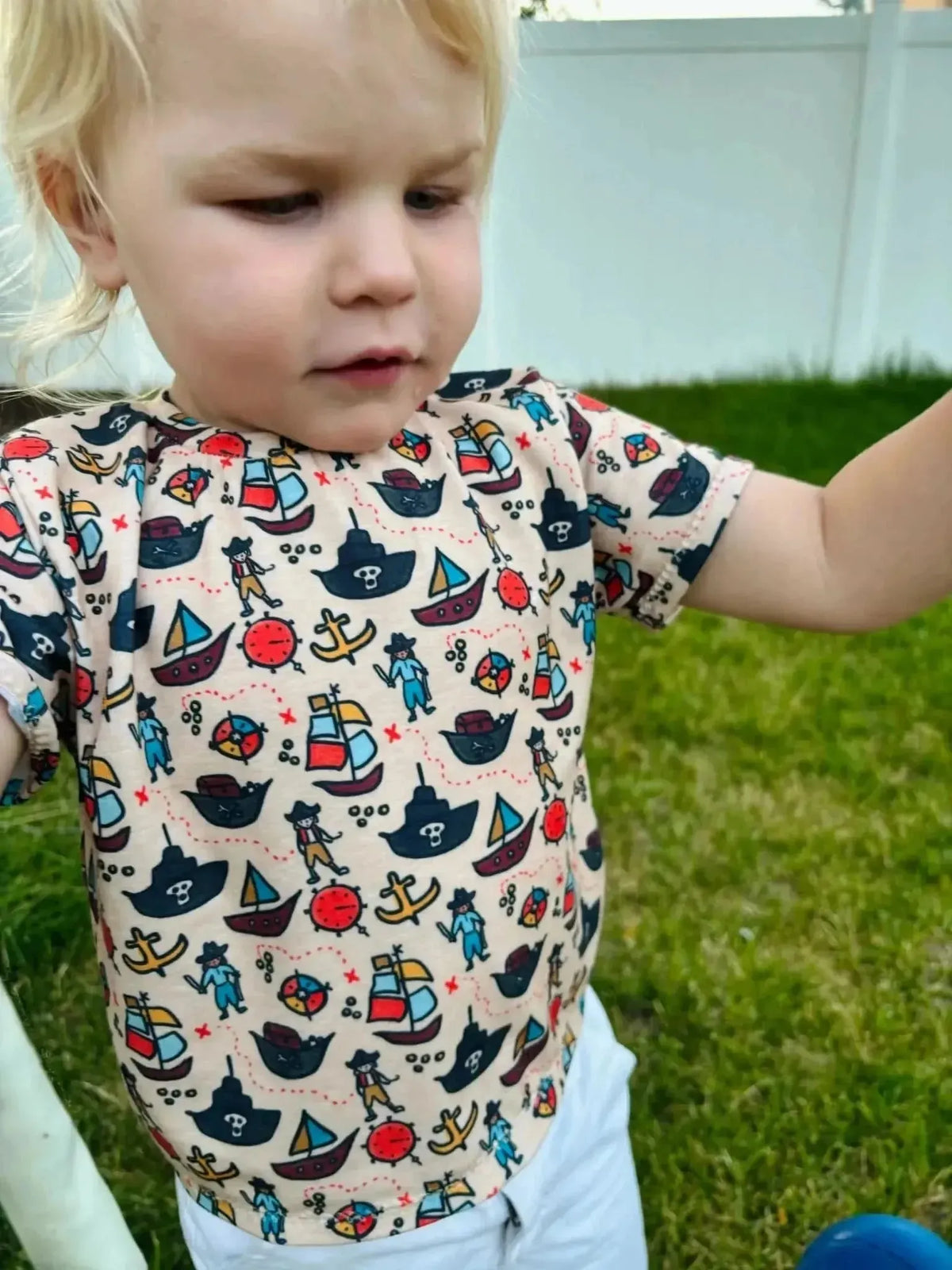 Blonde toddler wearing a short-sleeve shirt in WayMaker Fabrics Pirate Voyage print with ships and nautical motifs.