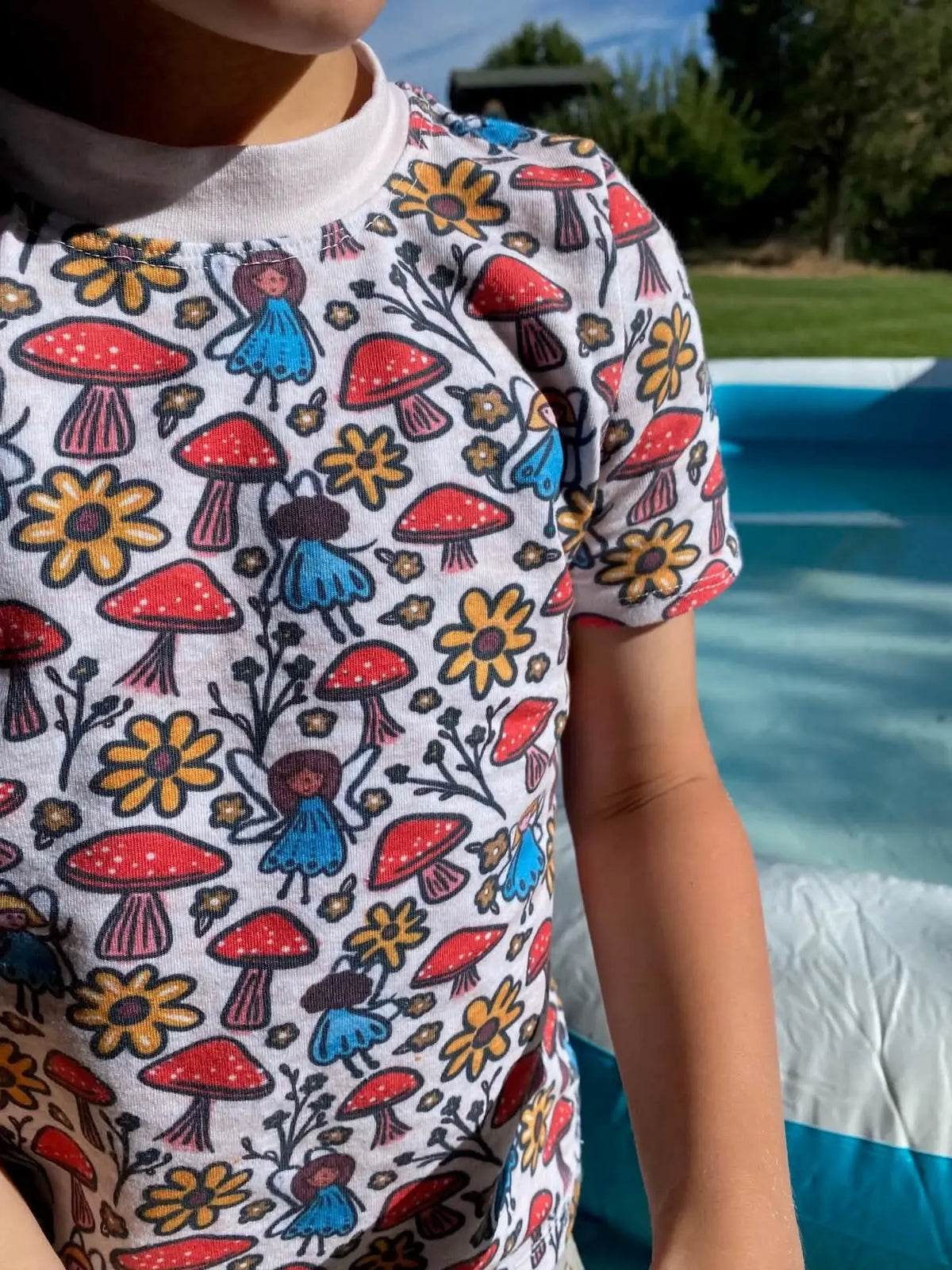 A child wears a white shirt with a pattern of mushrooms, flowers, and fairies.