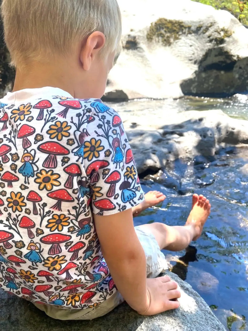 A child wears a white shirt with a colorful mushroom and fairy print.