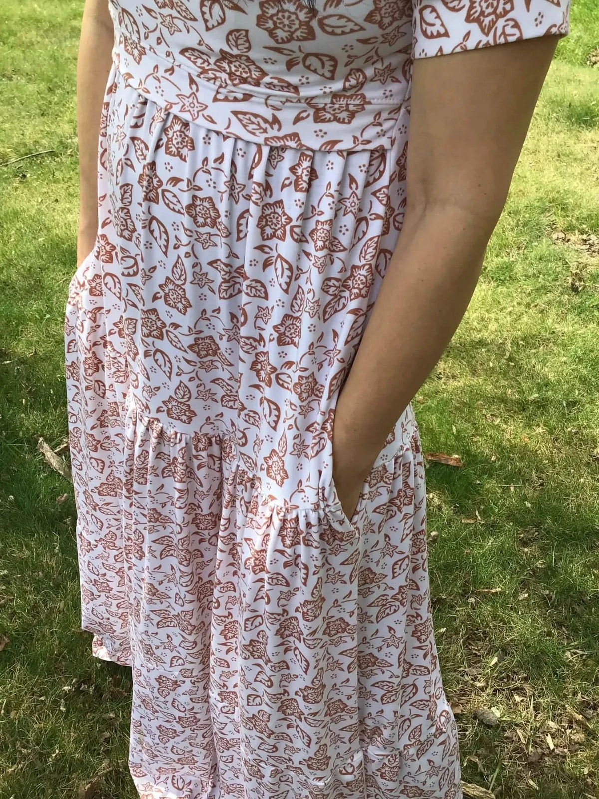 A person in a white dress with a brown floral pattern stands in green grass.