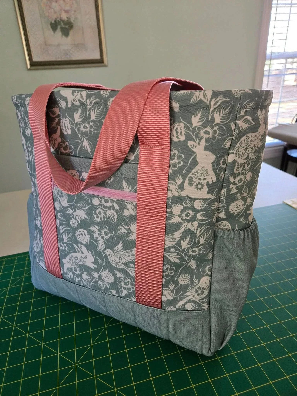 A tote bag with a sage green bunny and floral print, pink straps, and a denim base.