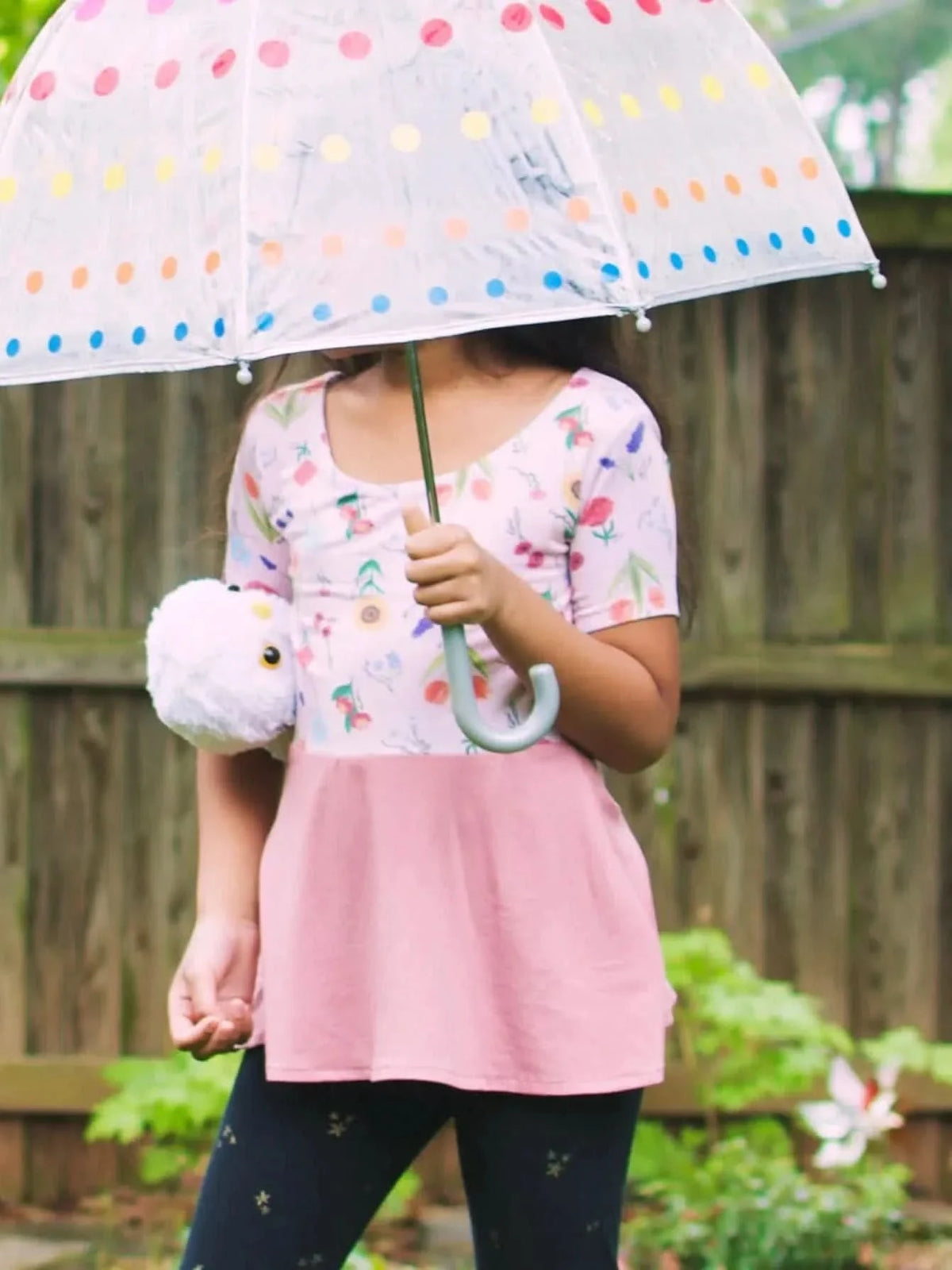 A child in a floral top and pink skirt holds a polka dot umbrella and white plush toy.