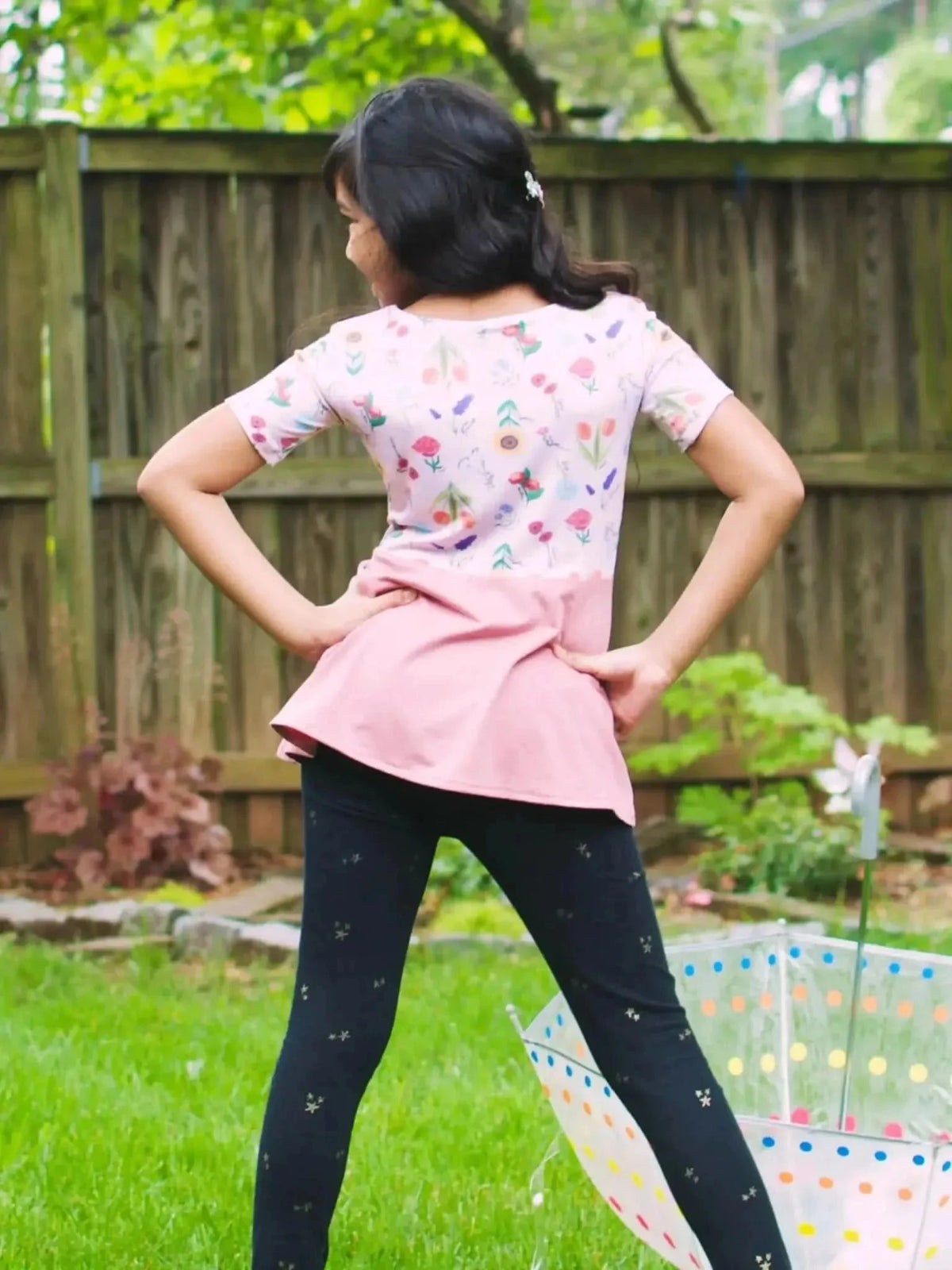 Girl wearing pink floral dress with butterfly garden print and black leggings in backyard.