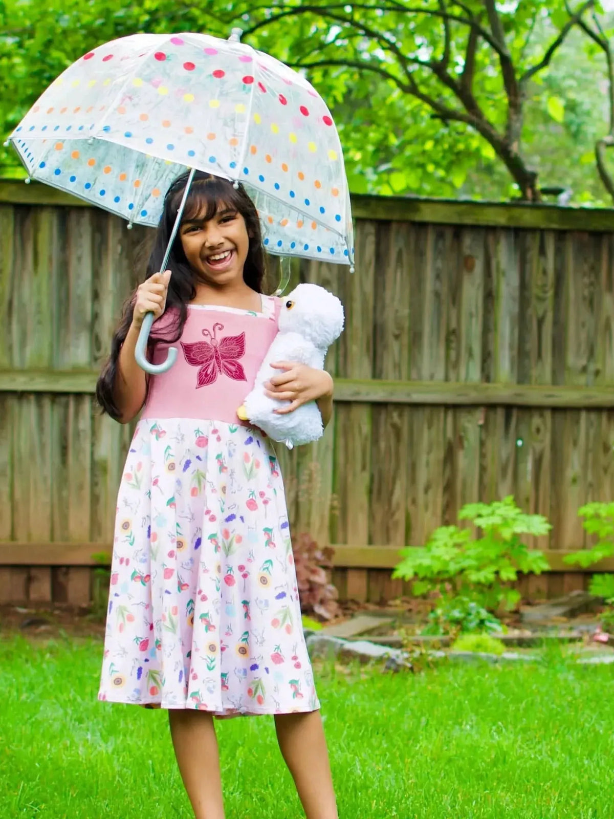 A young girl in a pink dress with a butterfly and floral skirt holds an umbrella.