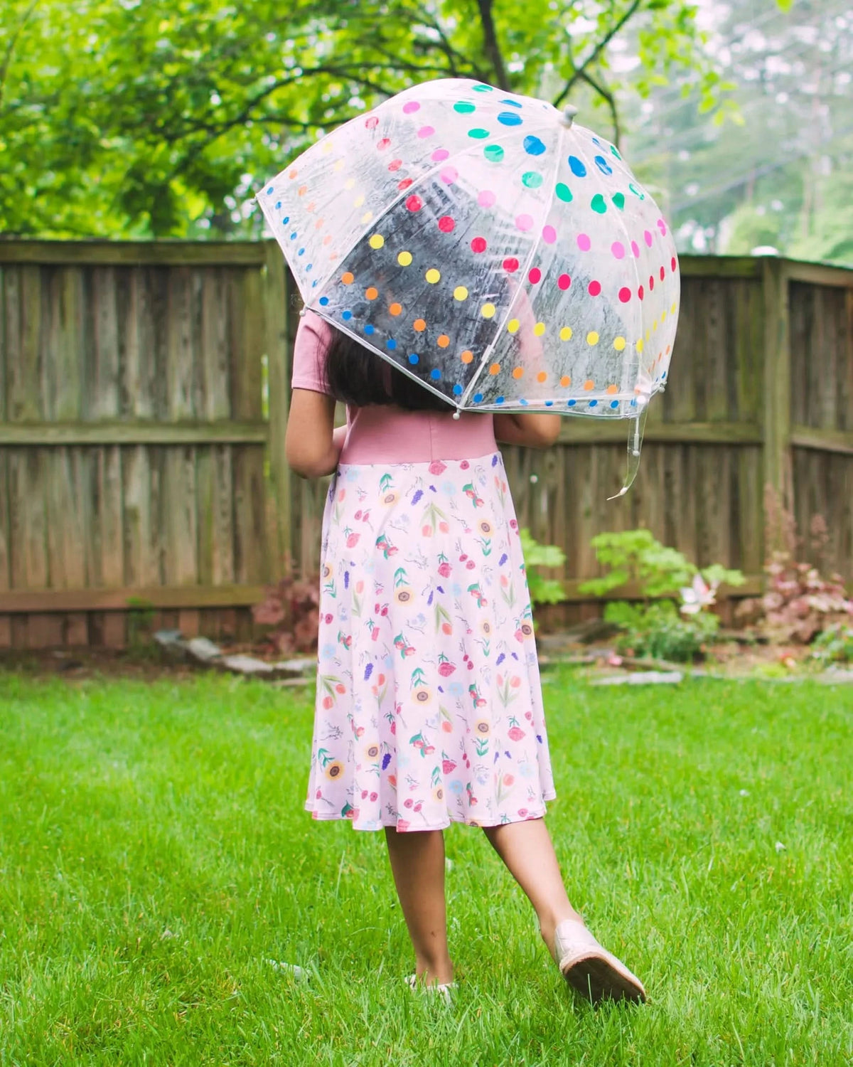 A person from behind holds a clear umbrella with colorful polka dots, wearing a floral skirt.
