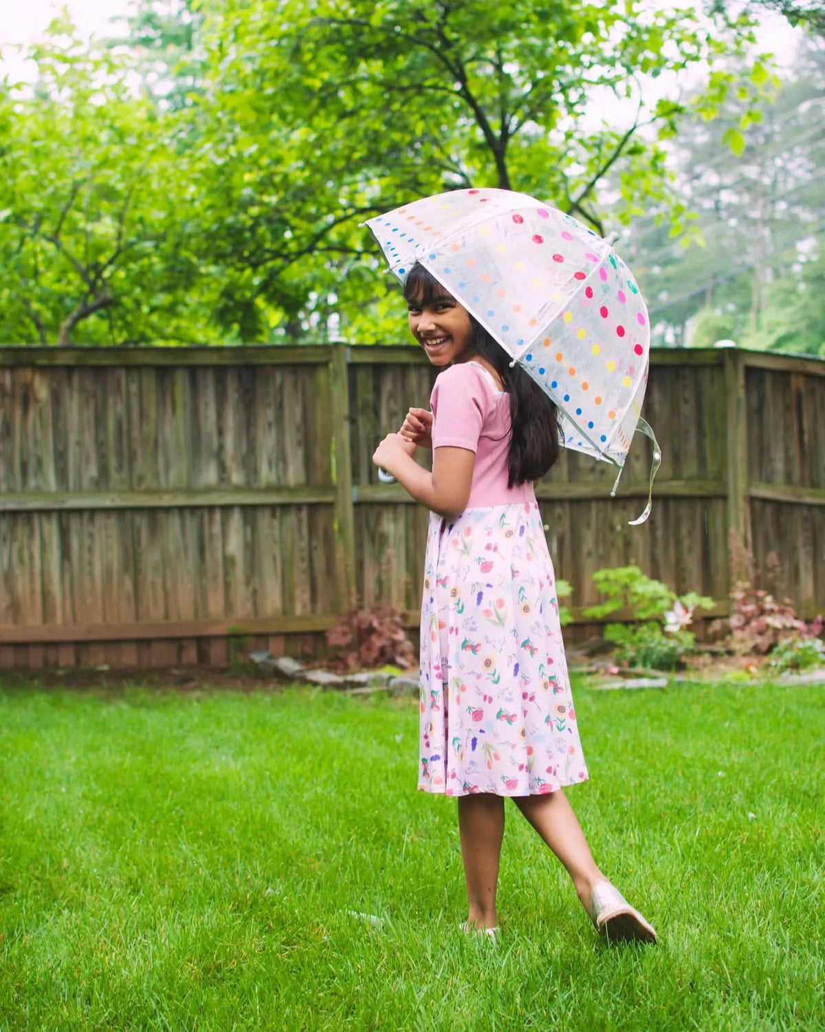 A young girl in a pink dress with a floral skirt holds a polka-dot umbrella.