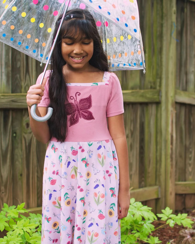 A girl in a pink dress with a butterfly and floral skirt holds a polka dot umbrella.