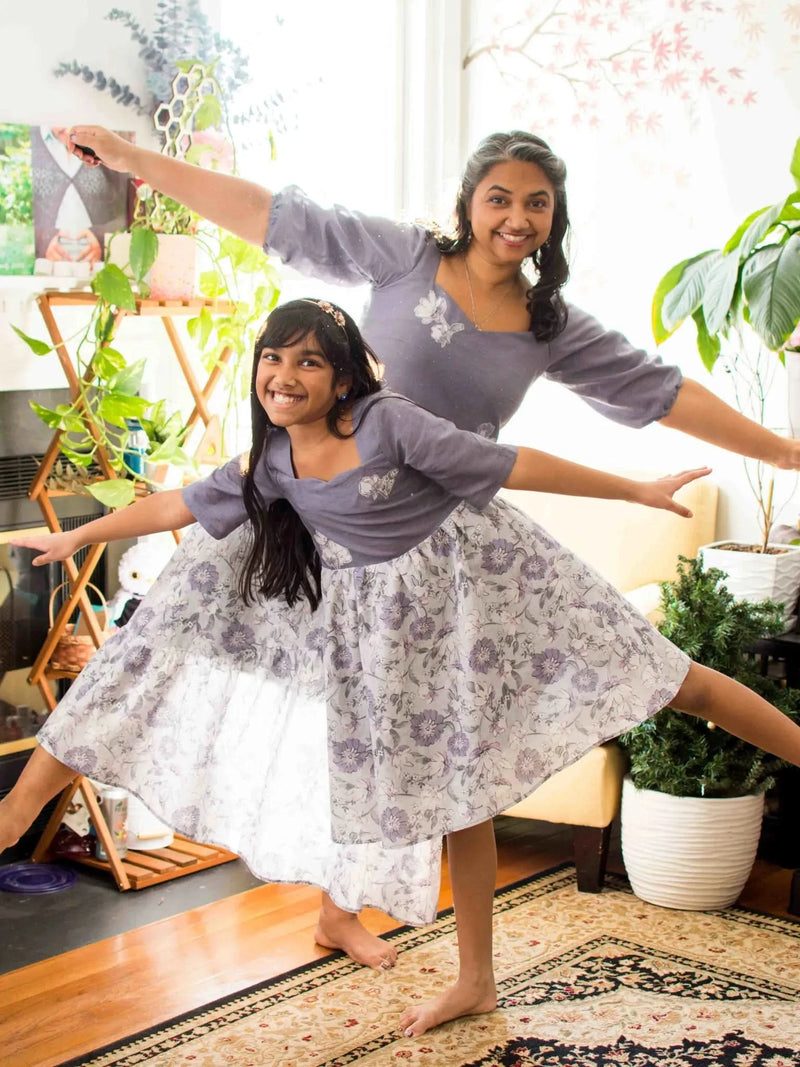 Two people in matching purple floral dresses pose with arms outstretched.