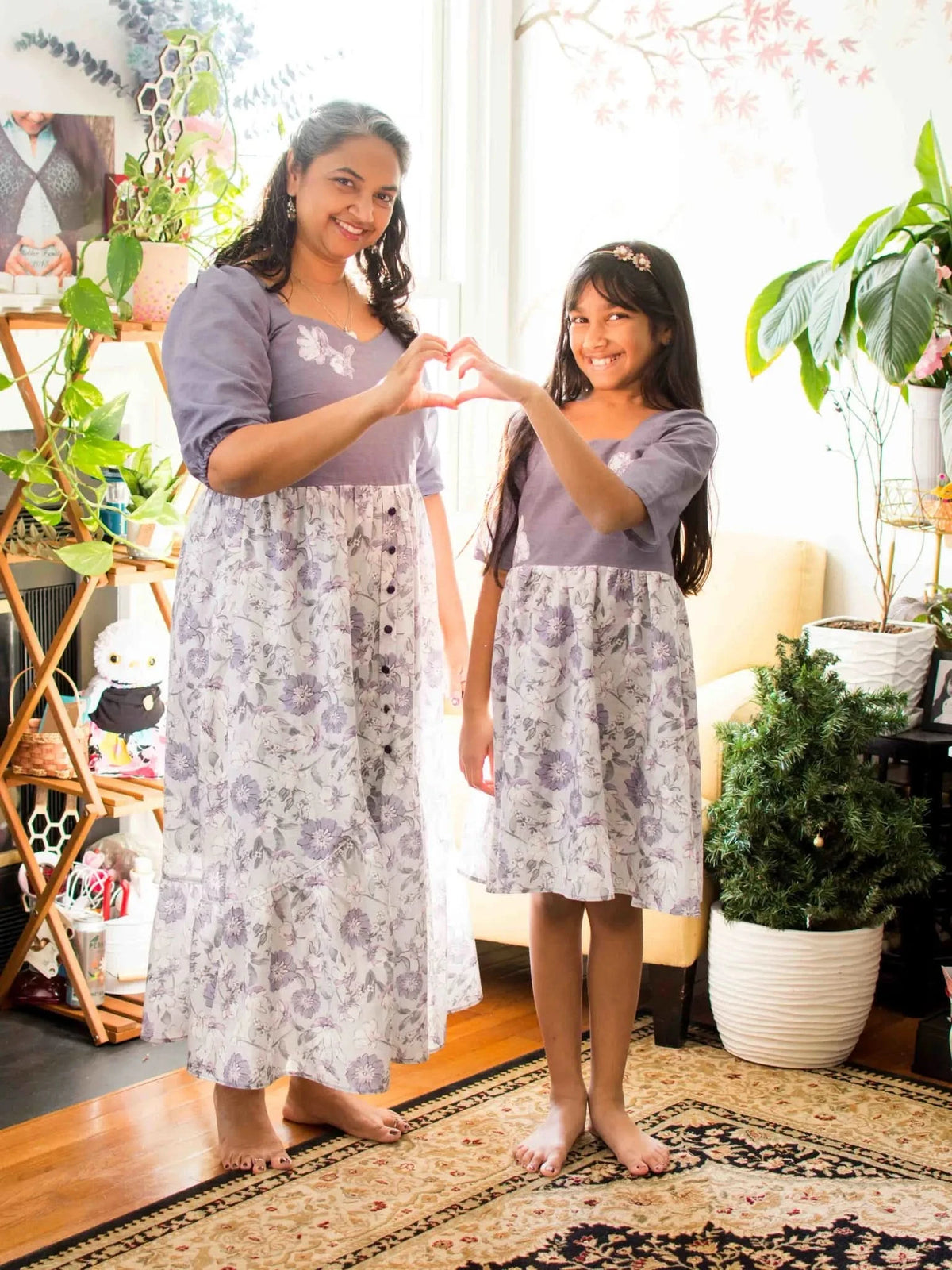 A mother and daughter in matching purple floral dresses form a heart with their hands.