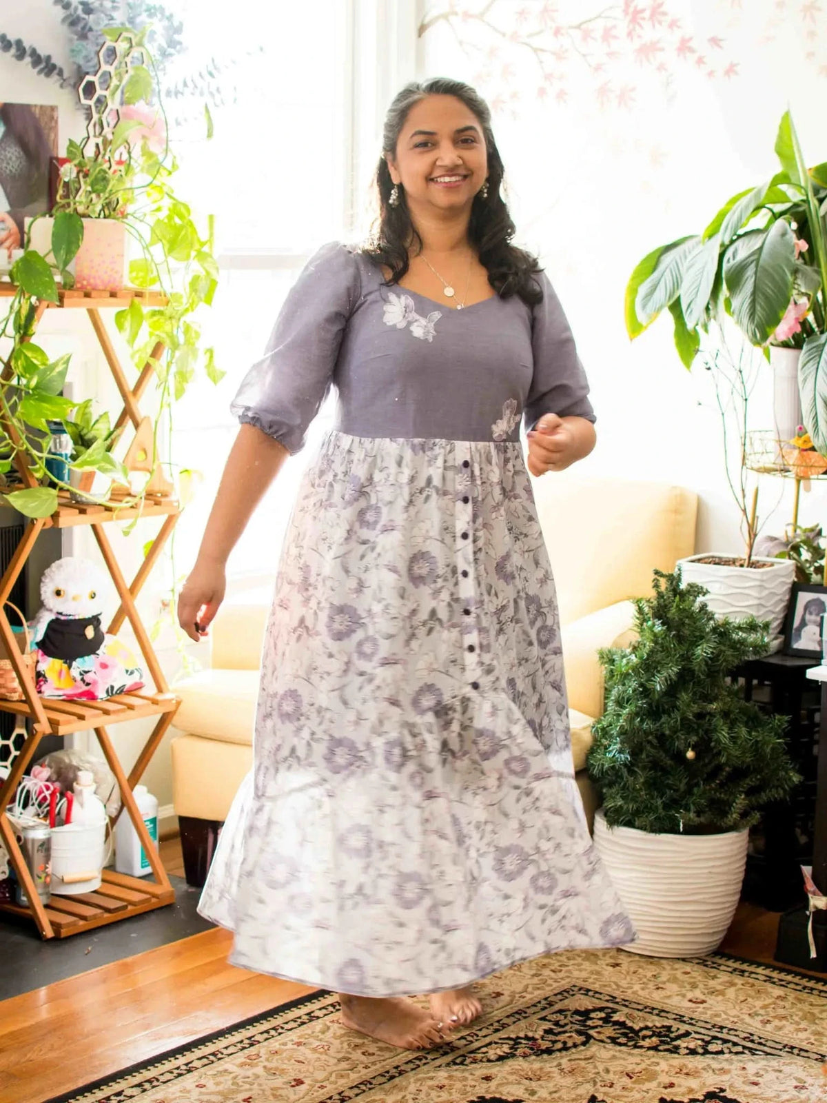 A woman in a purple floral dress smiles while standing on a patterned rug.