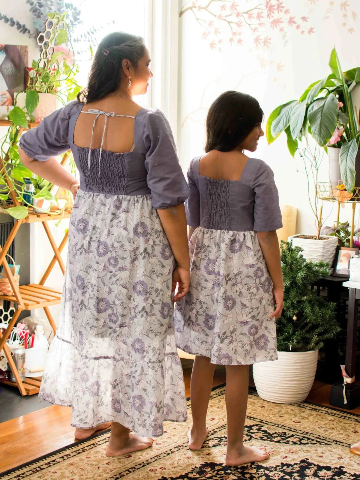 Two women from behind wear matching purple floral dresses with shirred bodices.