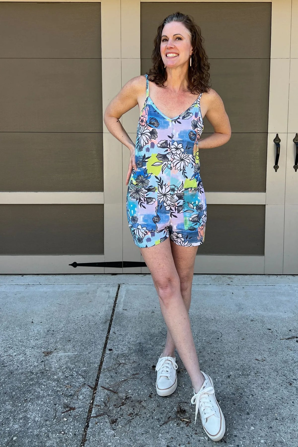 A woman smiles, wearing a blue graffiti floral romper and white sneakers.
