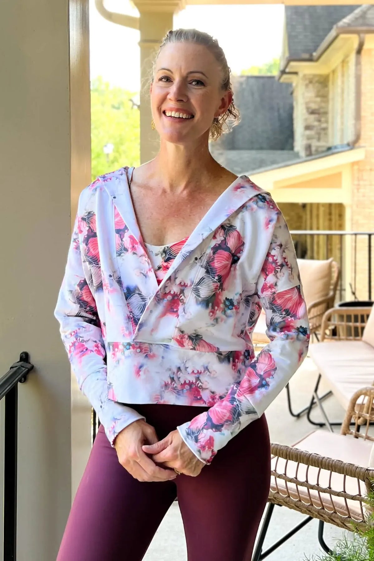 A woman smiles wearing a floral print wrap top and maroon leggings.