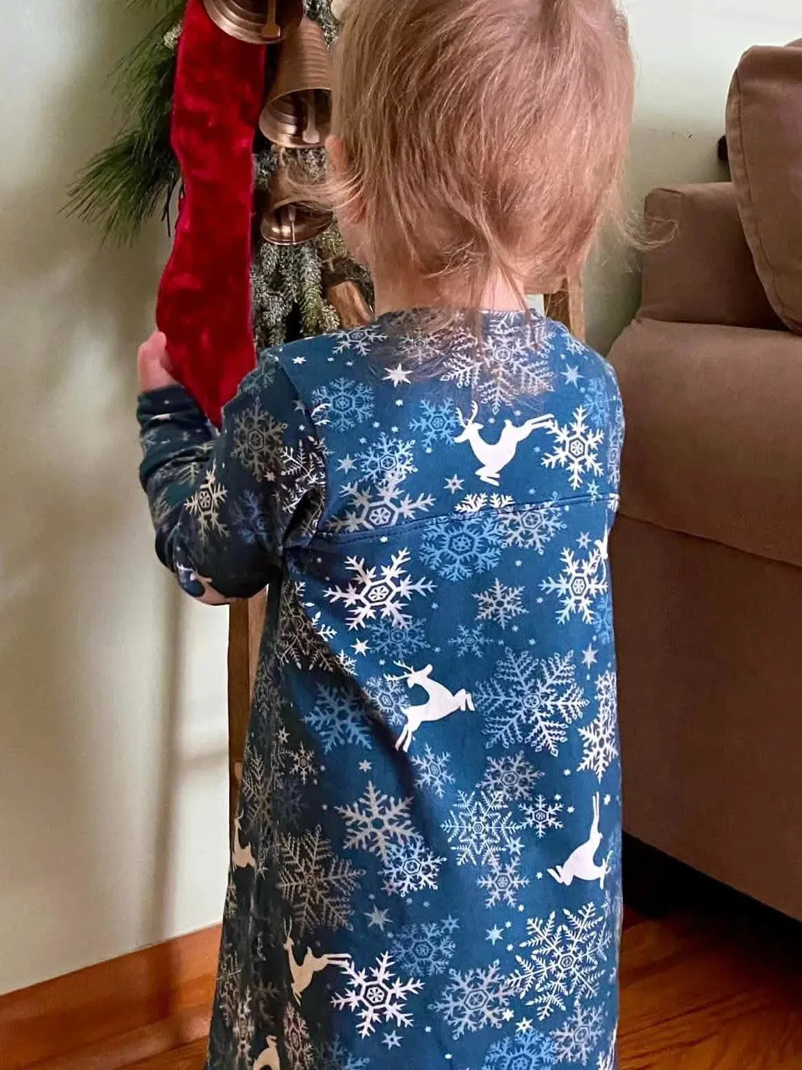 A child in a blue reindeer and snowflake print dress stands by a red stocking.