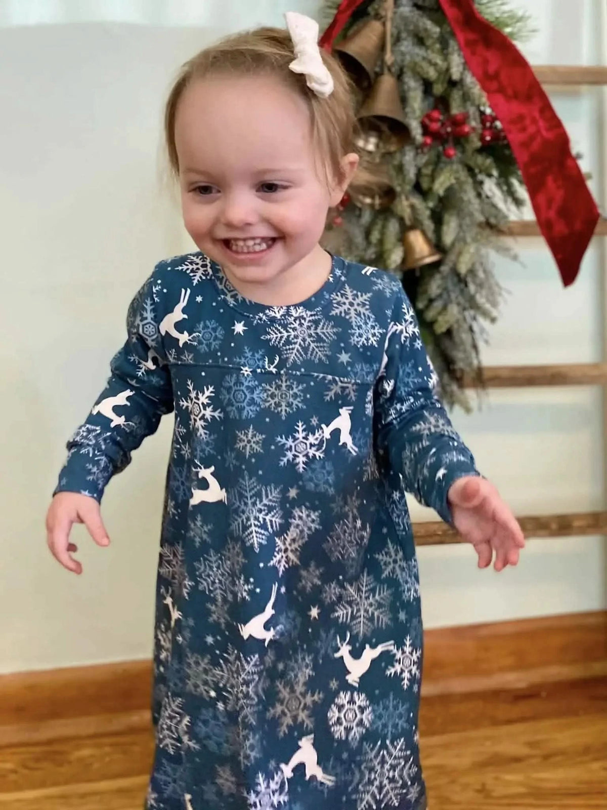 A smiling toddler in a blue dress with white reindeer and snowflake patterns.