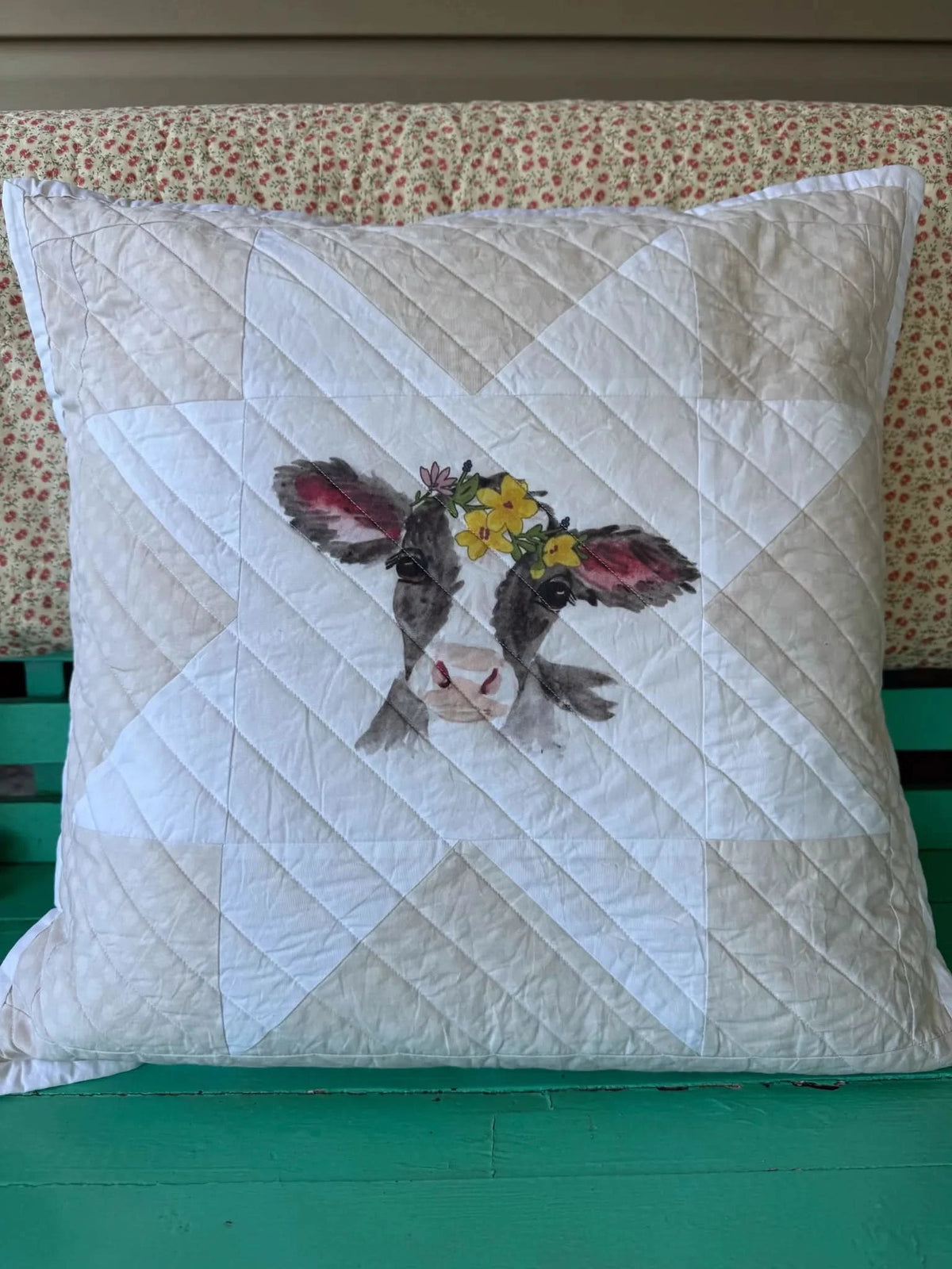 A quilted pillow with a cow wearing a flower crown, resting on a green bench.