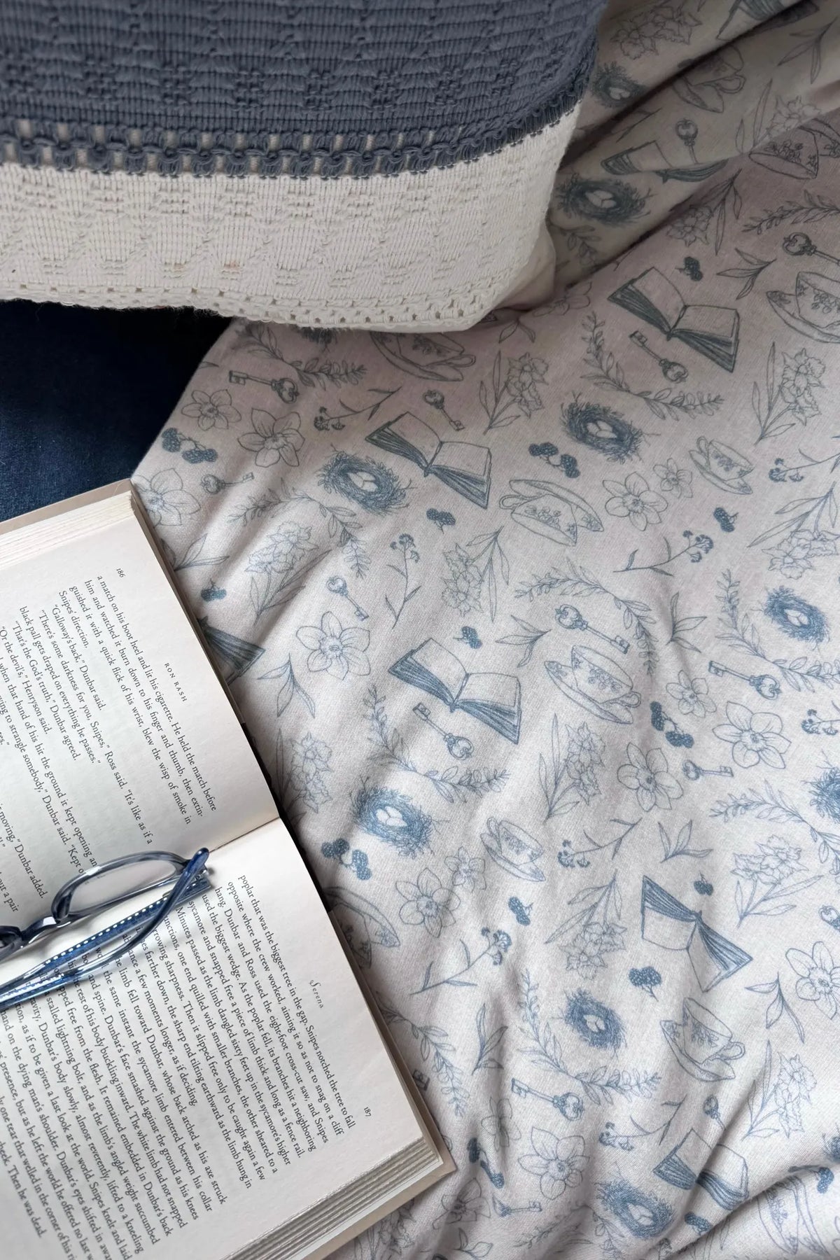 A book and glasses rest on a light fabric with a blue floral and object pattern.