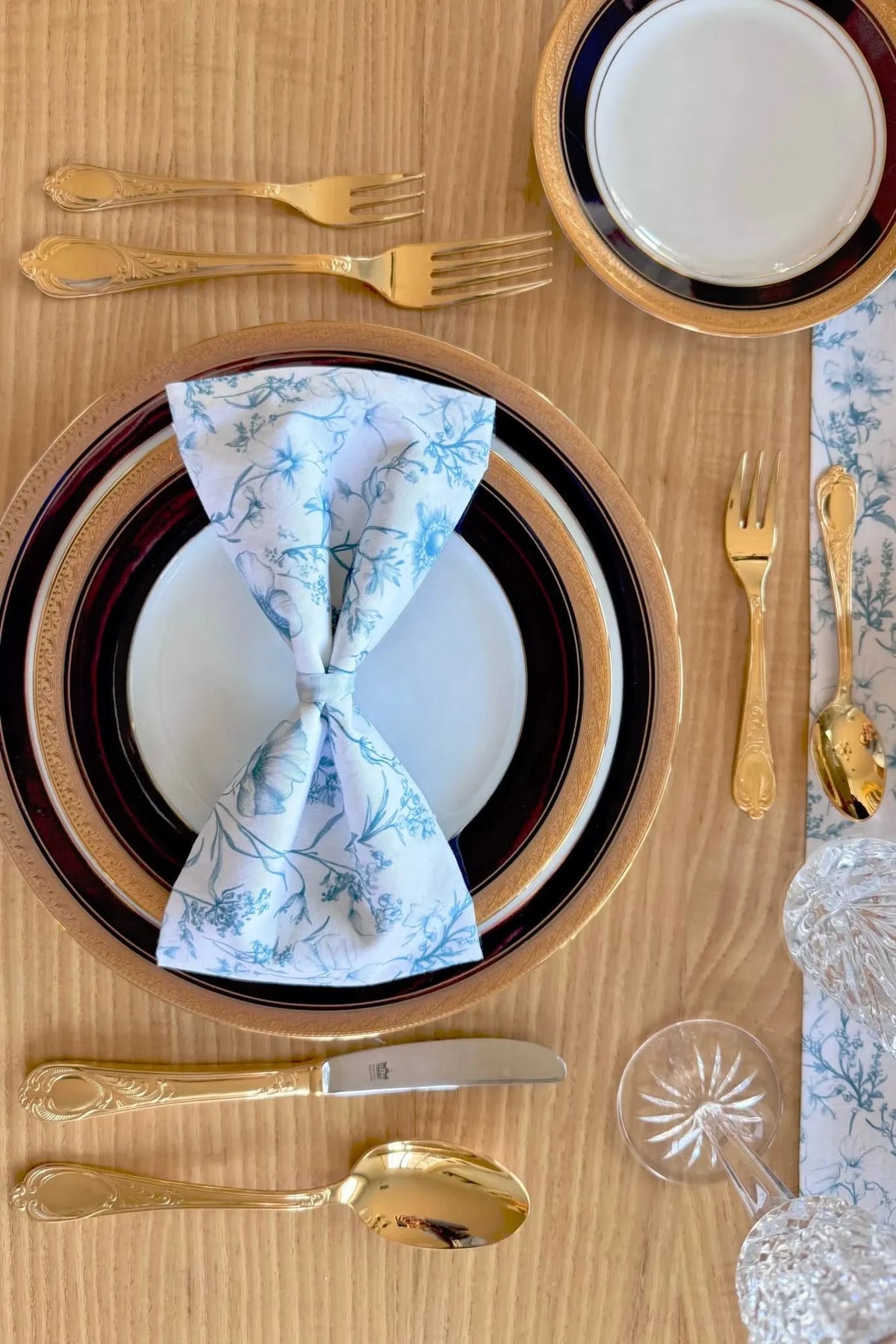 A formal place setting with gold flatware, ornate plates, and a blue floral napkin.