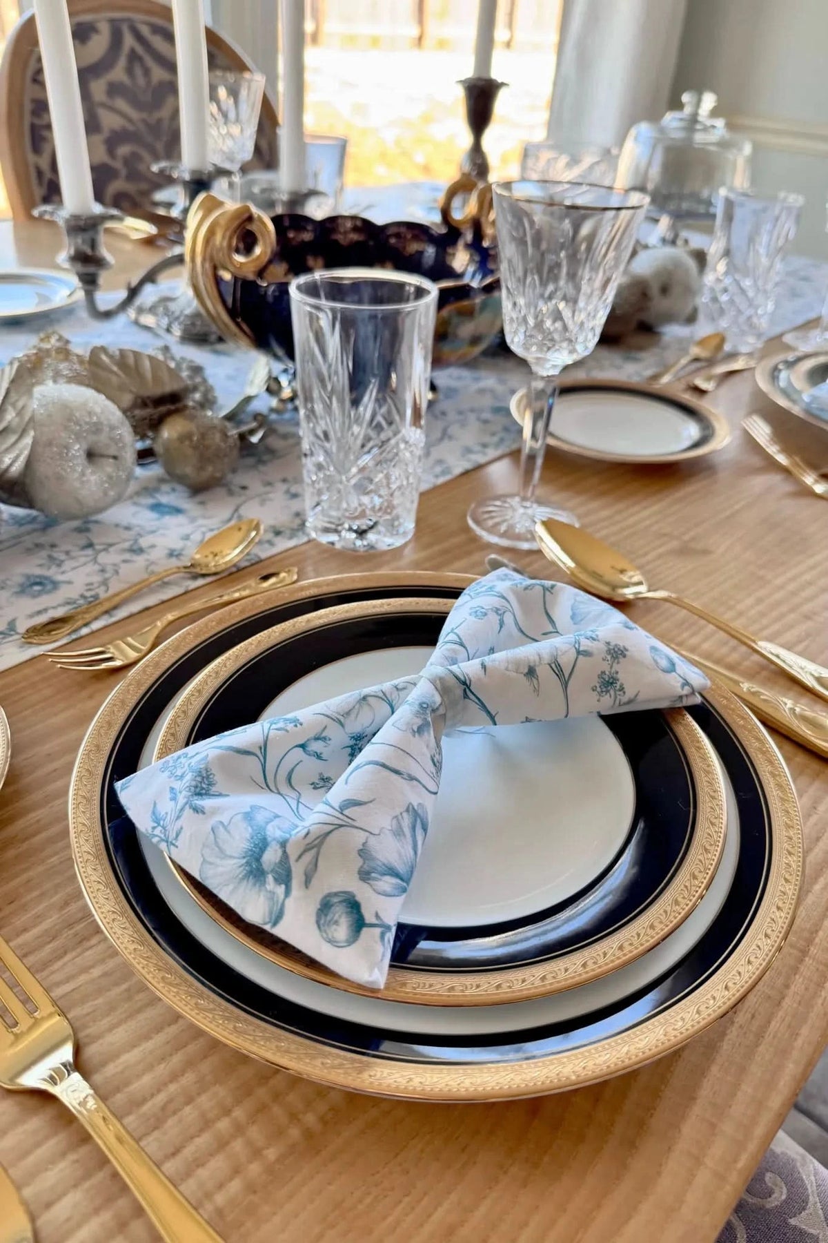A table set with blue and white floral napkins, gold-rimmed plates, and gold flatware.