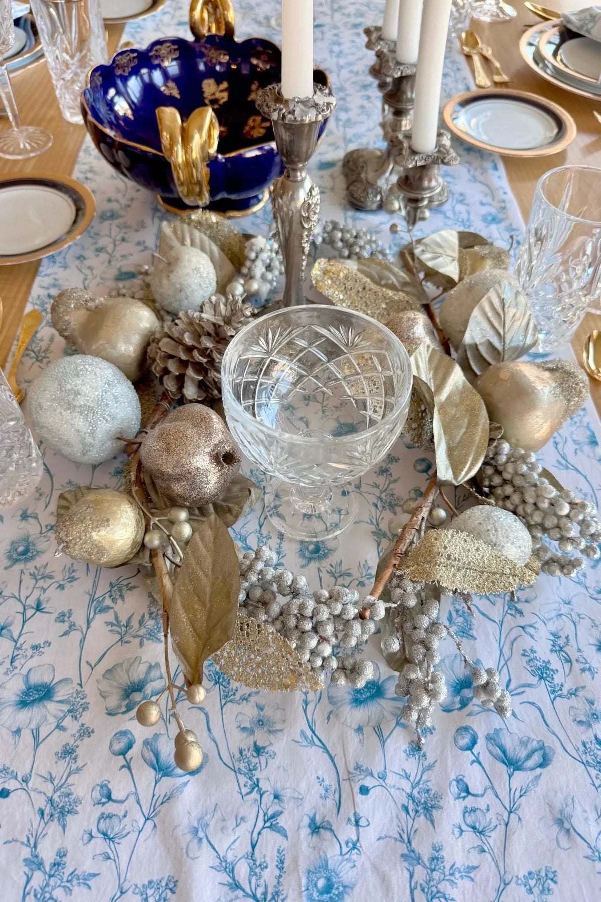 A table runner with a blue floral pattern is adorned with gold and silver holiday decorations.