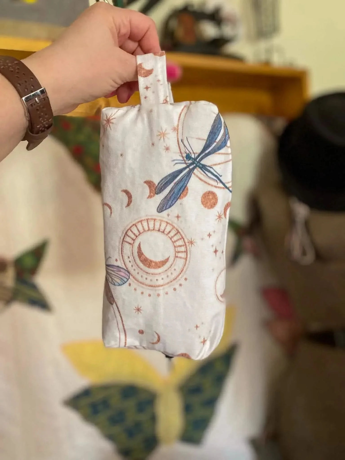 A hand holds a white fabric pouch with celestial dragonflies and moon phase patterns.