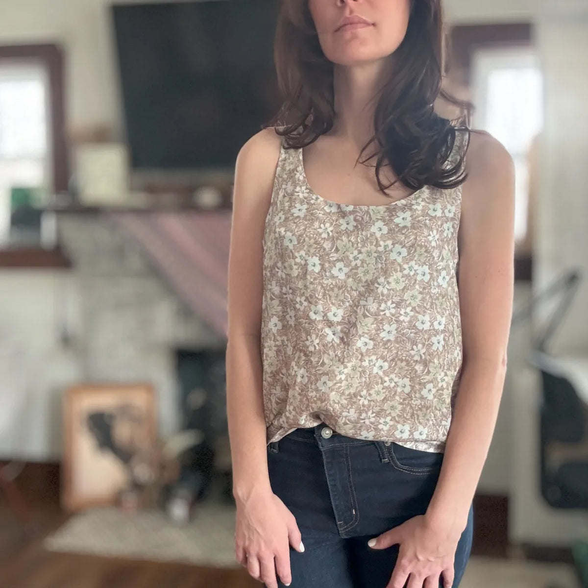 A woman wears a brown floral tank top and dark jeans.