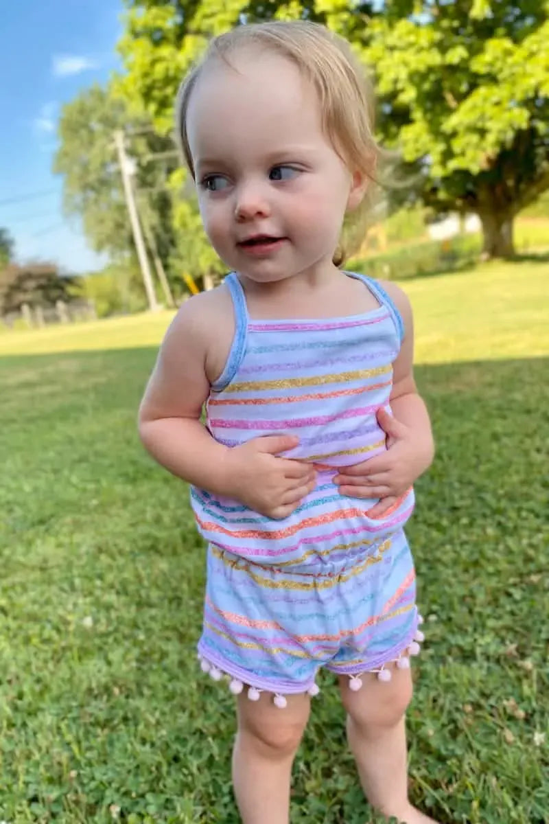 A toddler wears a striped romper with pom-pom trim, standing in grass.