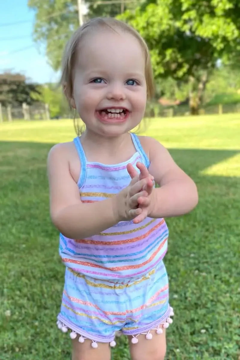 A toddler in a striped romper with pom-poms claps their hands in a grassy yard.