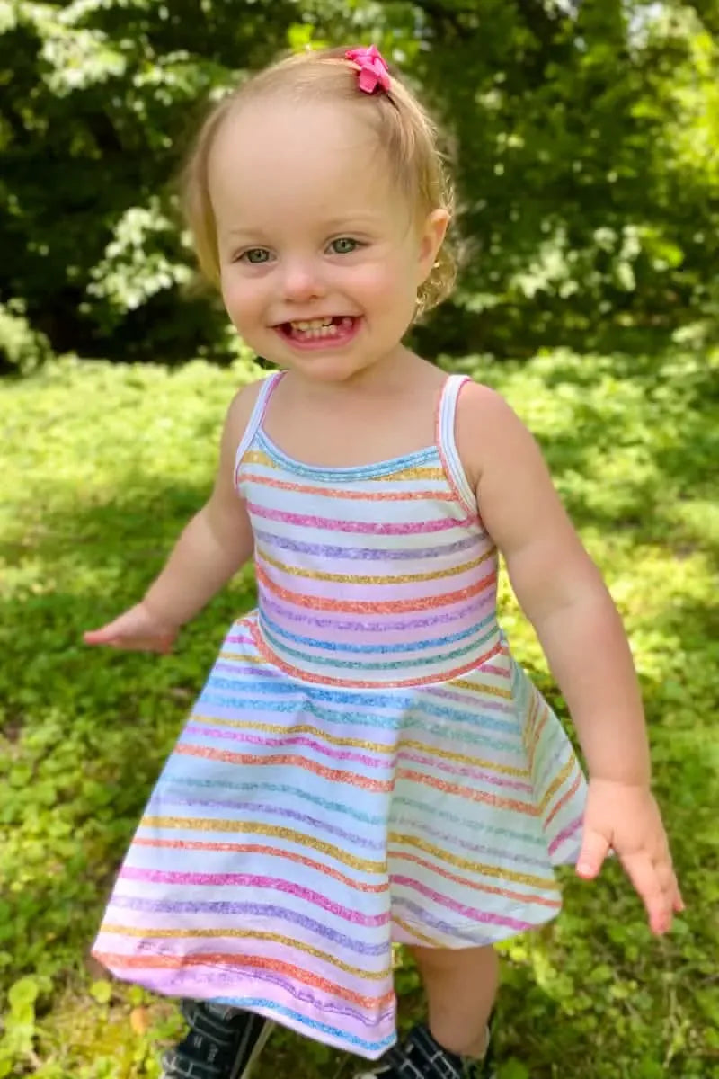 A smiling toddler wears a colorful striped dress with a pink bow in her hair.