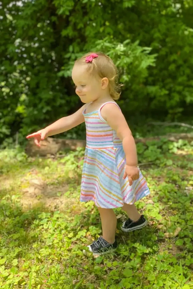 A toddler in a striped dress and black sneakers points to something in the grass.