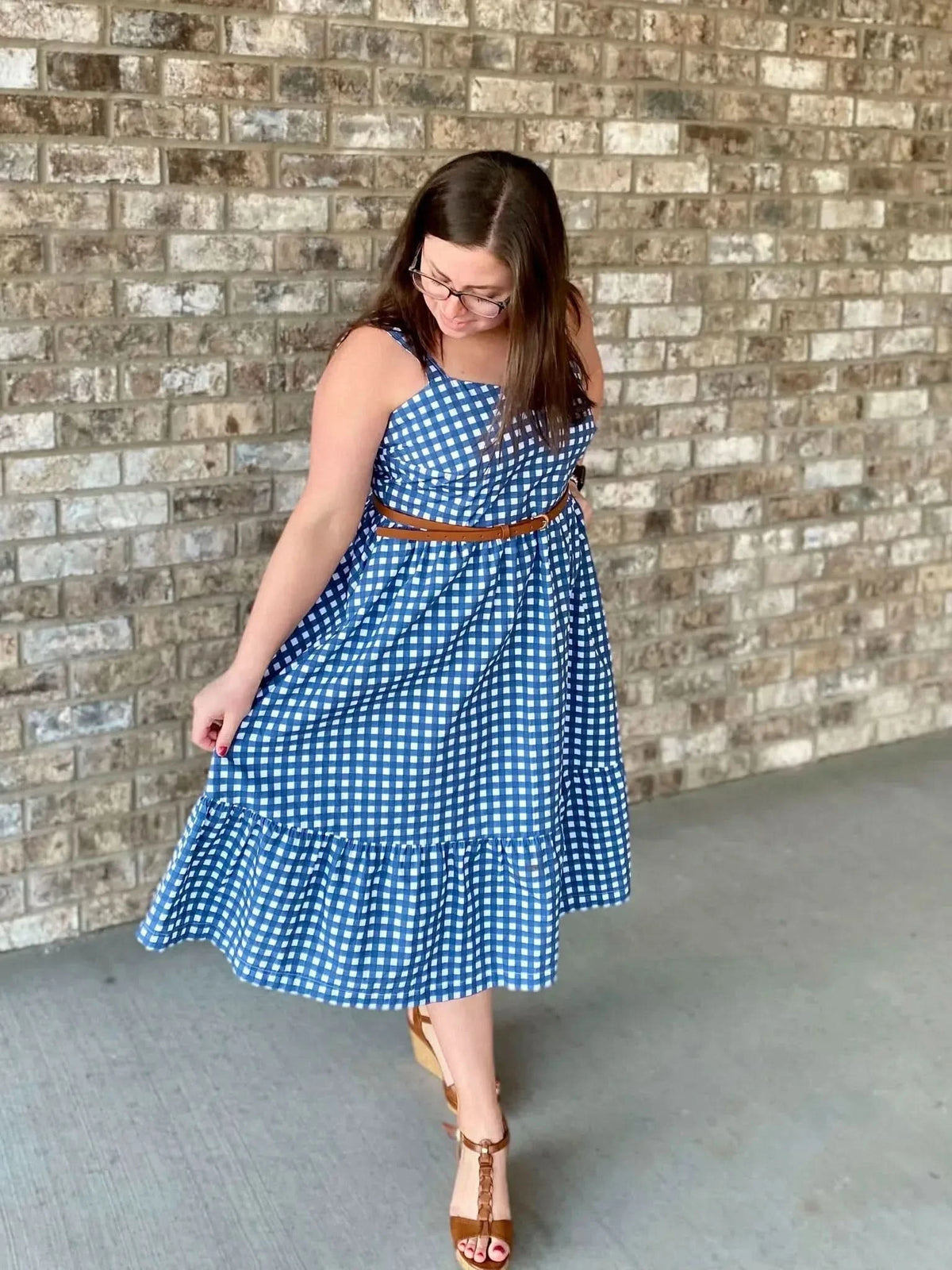 A woman in a navy gingham dress with a brown belt and sandals poses against a brick wall.
