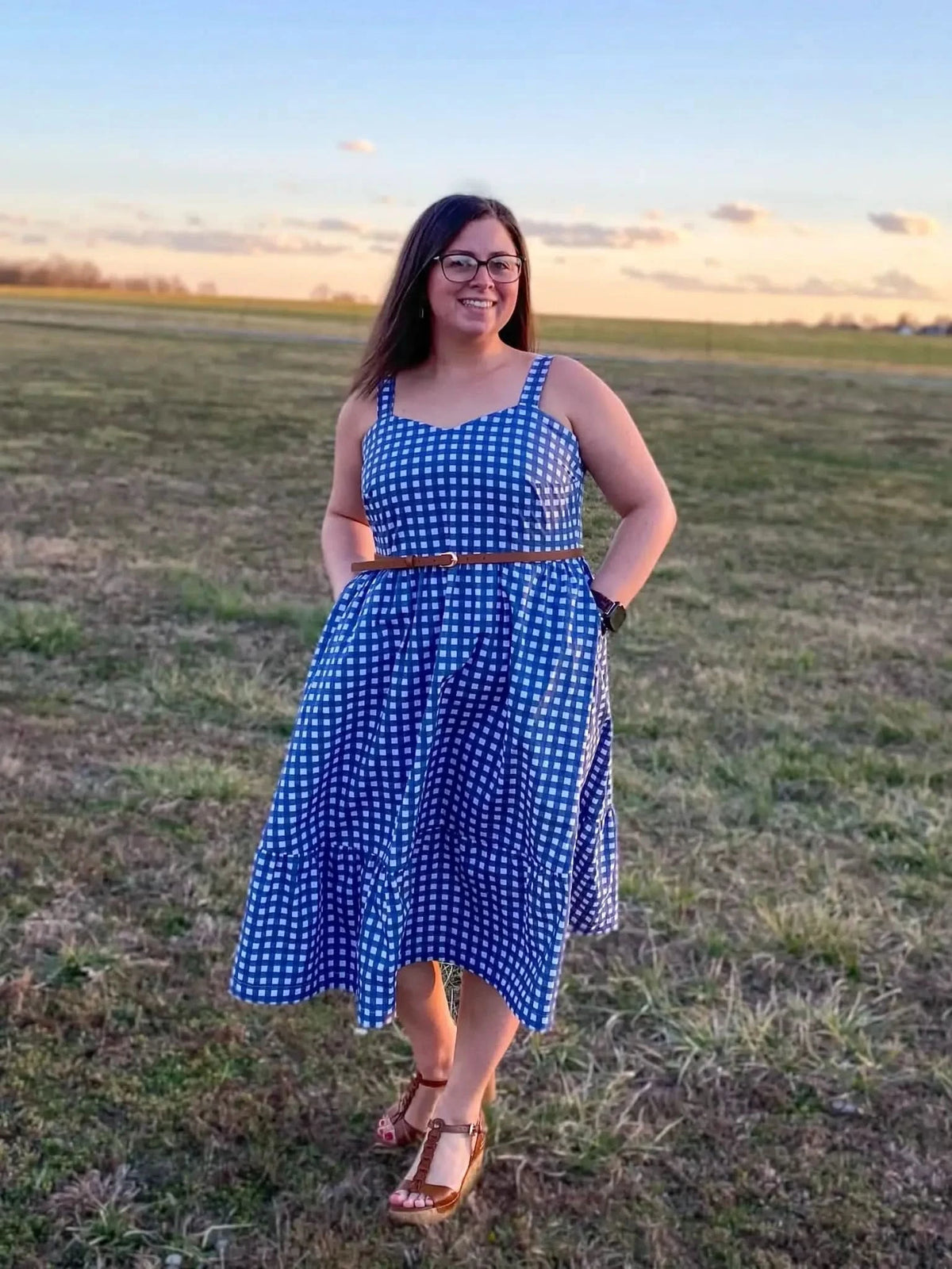 A woman in a blue and white gingham dress with a brown belt stands in a field.